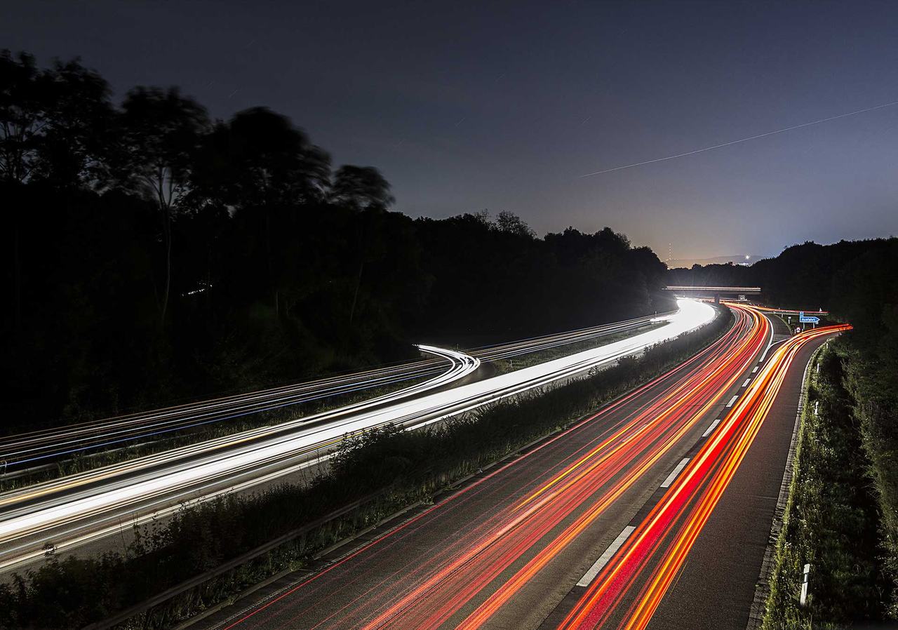 Autobahn bei Nacht: weiße Scheinwerfer- und rote Rücklicht-Lichtspuren ziehen in einer langen Kurve in die Ferne